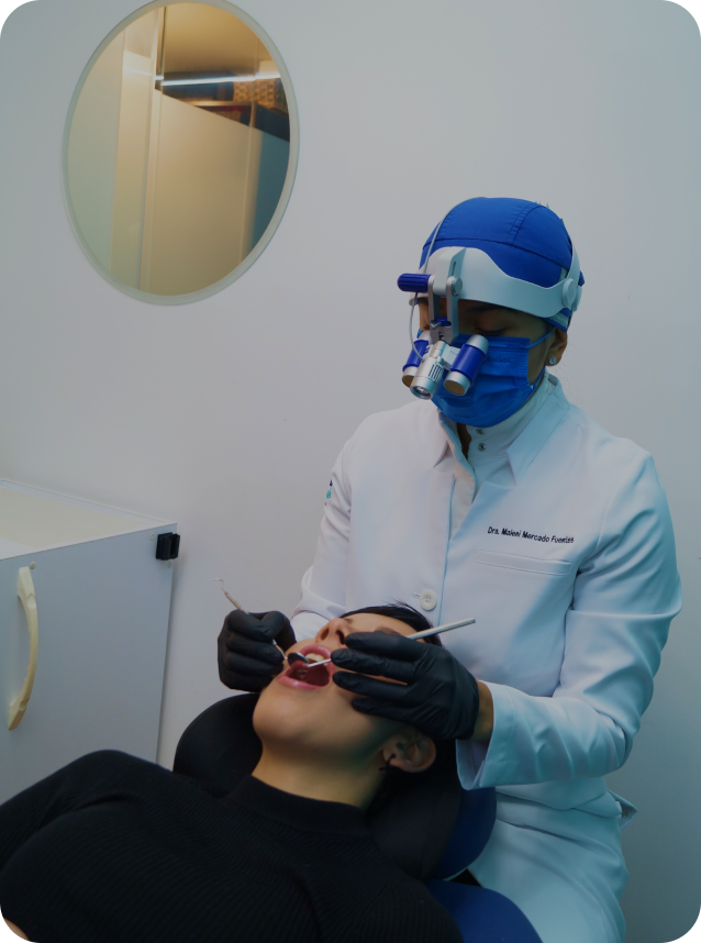 Dentista con bata blanca, gorro y lupa binocular realiza revisión dental a paciente acostado en silla odontológica.
