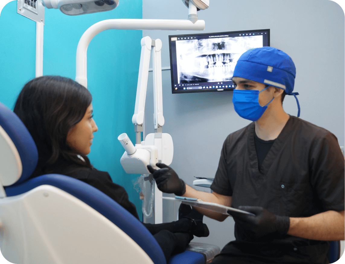 Dentista con gorro y cubrebocas azul revisa una radiografía dental mientras habla con paciente sentada en sillón dental.
